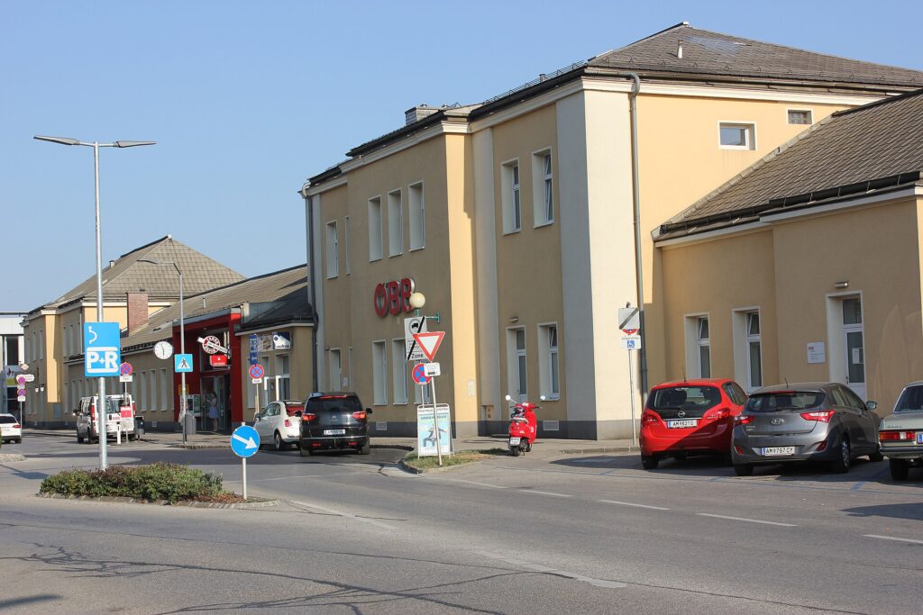 Schrägansicht des gelben, zweigeschoßigen Bahnhofsgebäudes von Amstetten mit rotem ÖBB-Logo, links niedrigere Flügelbauten mit rotem Eingangsbereich, davor ein Kreisverkehr mit Park-and-Ride-Schild und parkende Autos unter blauem Himmel