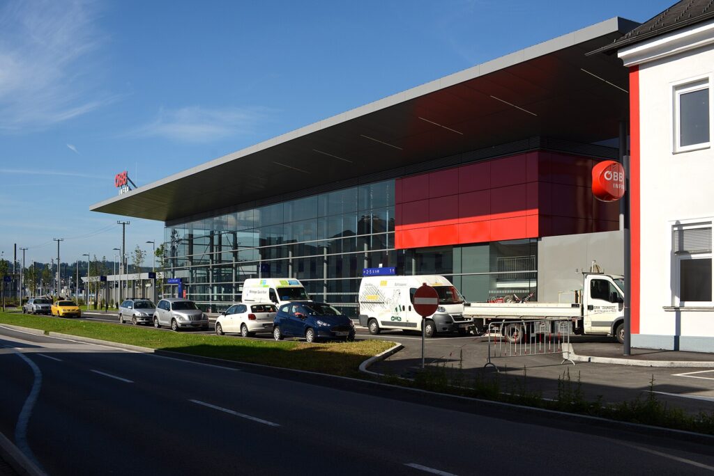 Die moderne Bahnhofshalle von Attnang-Puchheim mit weit auskragendem Flachdach, Glasfassade und roter Wandfläche, davor geparkte Autos und Lieferwagen an der Straße unter blauem Himmel