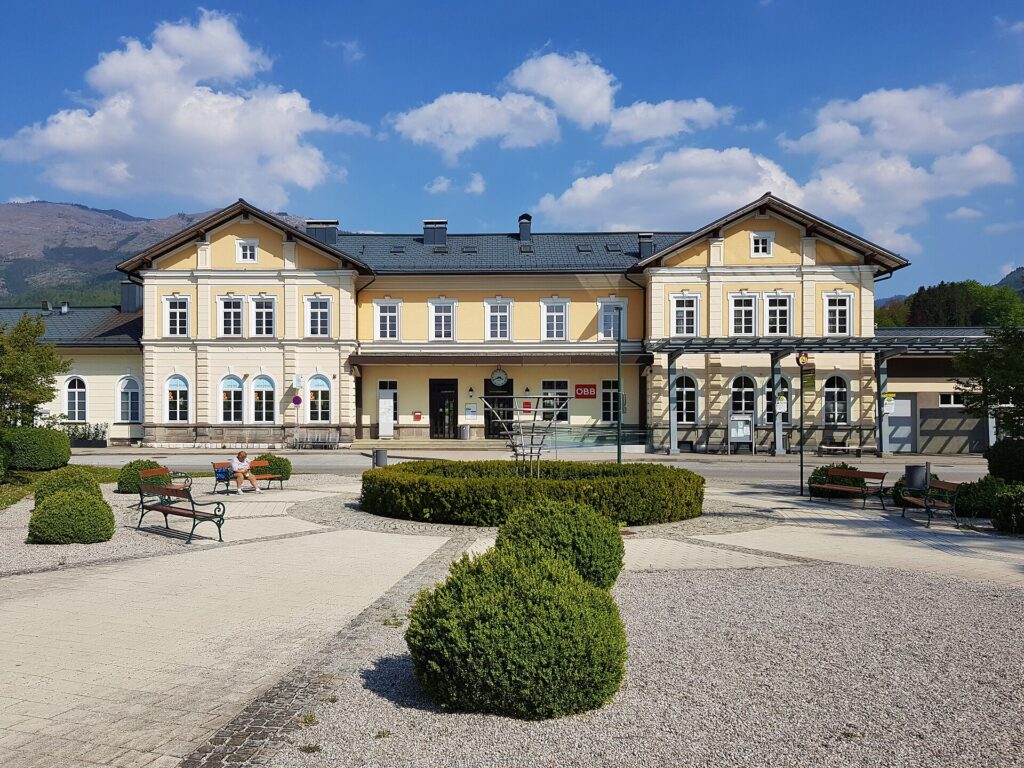 Frontansicht des gelb-weißen historistischen Aufnahmsgebäudes von Bad Ischl mit zwei Seitenrisaliten mit Giebeln, davor ein gepflasterter Platz mit Buchskugeln und Parkbänken unter blauem Himmel mit Wolken