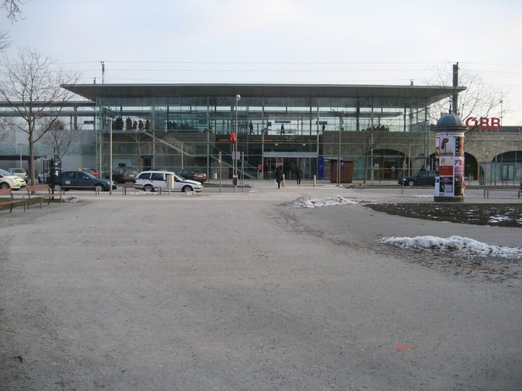 Das flache Bahnhofsgebäude von Baden aus Glas und Stahl mit weit auskragendem Dach, von vorne über den leeren Vorplatz mit Schneeresten gesehen, links geparkte Autos, rechts eine Litfaßsäule