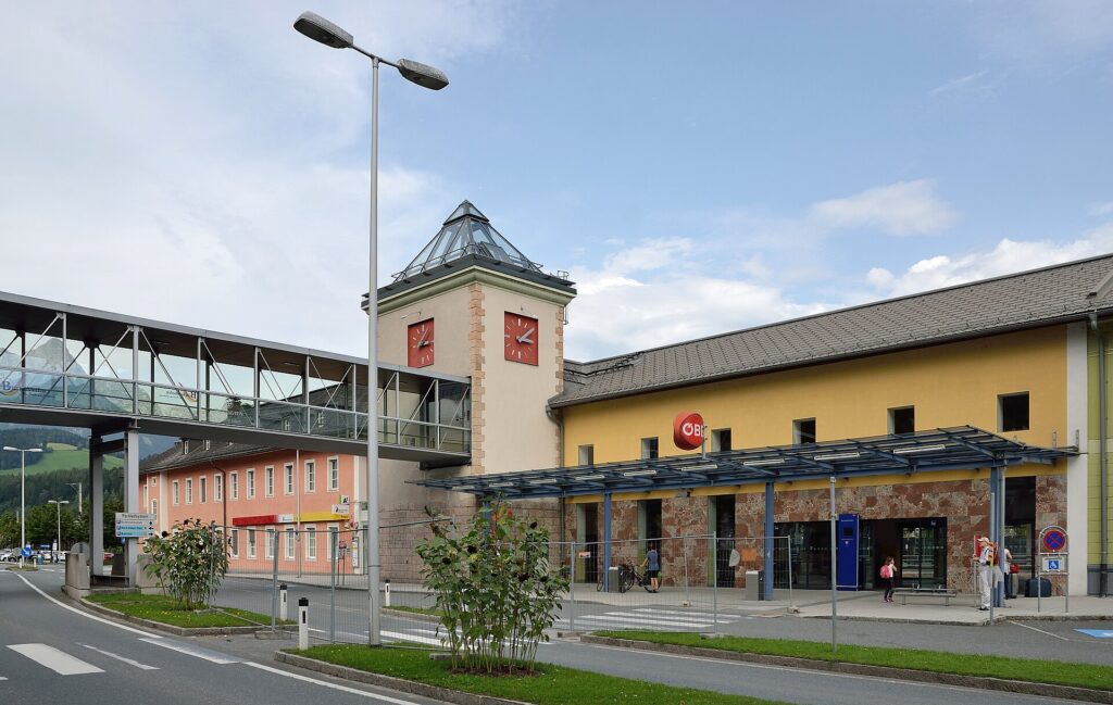 Straßenseitige Ansicht des gelben Aufnahmsgebäudes von Bischofshofen mit ÖBB-Logo, Glasvordach und Uhrturm mit Glaspyramide, links ein verglaster Fußgängersteg und ein rosa Nachbargebäude unter blauem Himmel