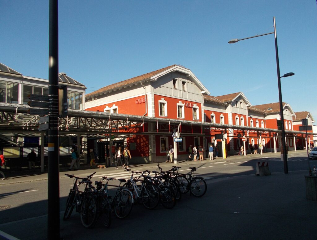 Schrägansicht des zweigeschoßigen, rot gestrichenen Bahnhofsgebäudes mit drei Quergiebeln, ÖBB-Schriftzug und langem Vordach über dem Gehweg, links die Glas-Stahl-Überdachung des Haupteingangs, im Vordergrund abgestellte Fahrräder unter blauem Himmel
