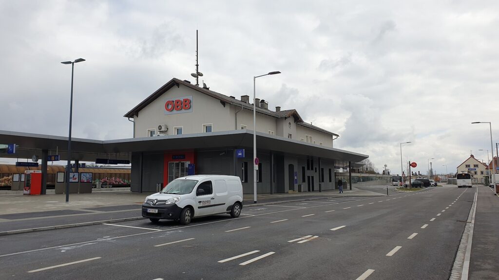 Das zweigeschoßige helle Aufnahmsgebäude von Braunau am Inn mit ÖBB-Schriftzug und langem Vordach am Busvorplatz, davor ein weißer Lieferwagen unter bewölktem Himmel