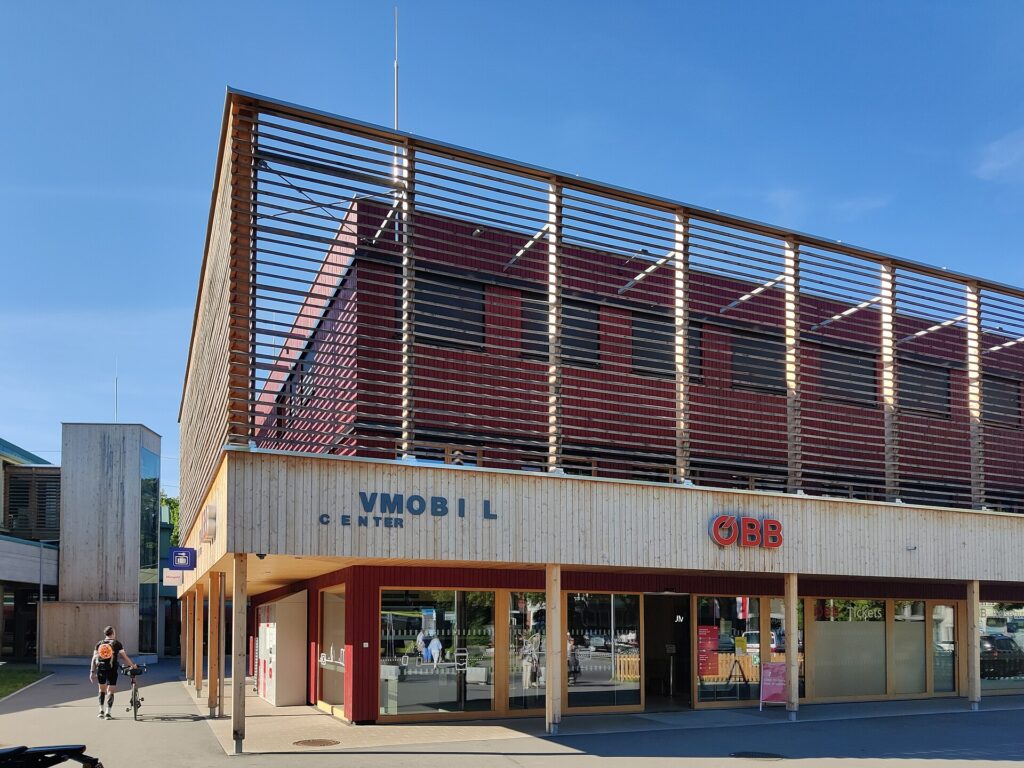 Das dreistöckige Interimsgebäude des Bahnhofs Bregenz aus Holz mit roter Fassade hinter waagrechten Holzlamellen, im Erdgeschoß große Glasfenster mit den Schriftzügen VMOBIL Center und ÖBB, links ein hölzerner Liftturm, davor ein Radfahrer