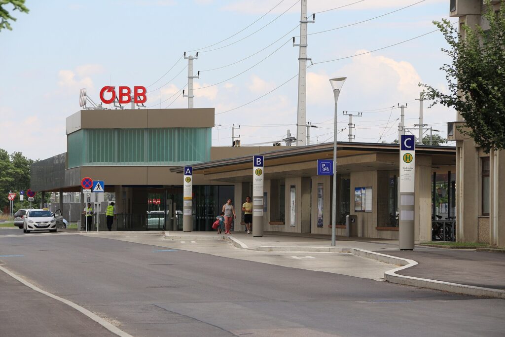 Das neue Bahnhofsgebäude von Bruck an der Leitha mit grün verglastem Aufbau und rotem ÖBB-Logo, davor das lange Vordach mit den Haltestellensäulen der Bussteige A, B und C am Bahnhofplatz