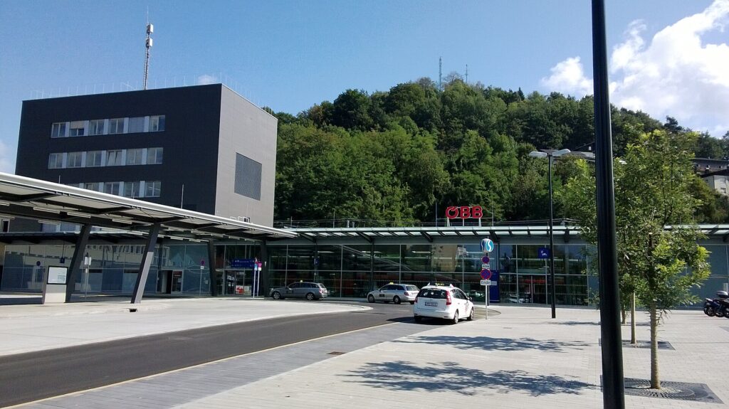 Der Vorplatz des Bahnhofs Bruck an der Mur mit überdachten Bussteigen, dahinter der dunkle mehrgeschoßige Turm und die verglaste Bahnhofshalle mit rotem ÖBB-Schriftzug vor einem bewaldeten Hang, davor Taxis