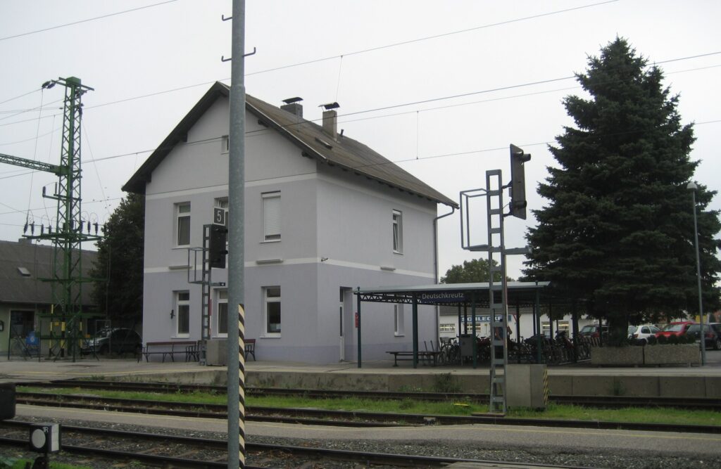 Das zweigeschoßige, hellgrau-fliederfarbene Bahnhofsgebäude mit Satteldach hinter den Gleisen, daneben ein grünes Bahnsteigdach mit dem Ortsschild Deutschkreutz, Fahrradständer und eine große Fichte