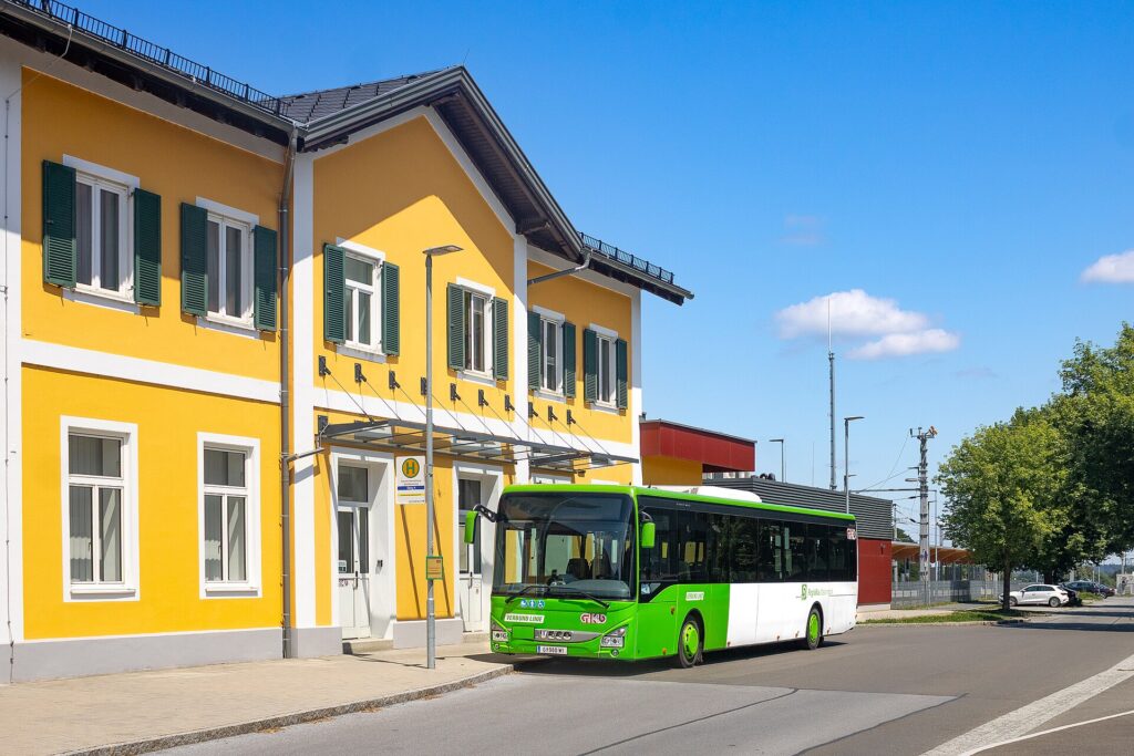 Das zweigeschoßige gelbe Bahnhofsgebäude von Deutschlandsberg mit weißen Fensterrahmen und grünen Fensterläden, davor ein grün-weißer GKB-Regionalbus an der Haltestelle unter blauem Himmel