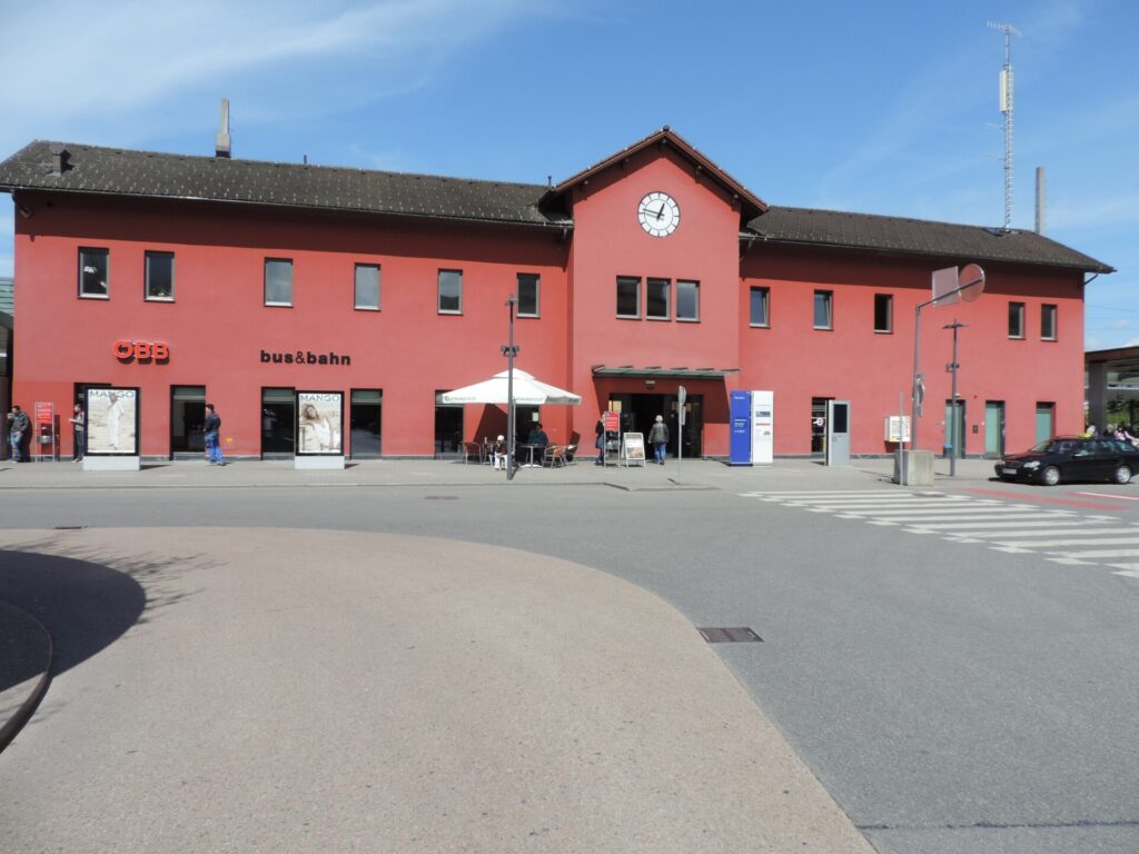 Frontalansicht des roten zweigeschoßigen Bahnhofsgebäudes von Dornbirn mit Satteldach und Uhr im Giebel, an der Fassade die Schriftzüge ÖBB und bus&bahn, davor die Straße am Bahnhofsvorplatz unter blauem Himmel