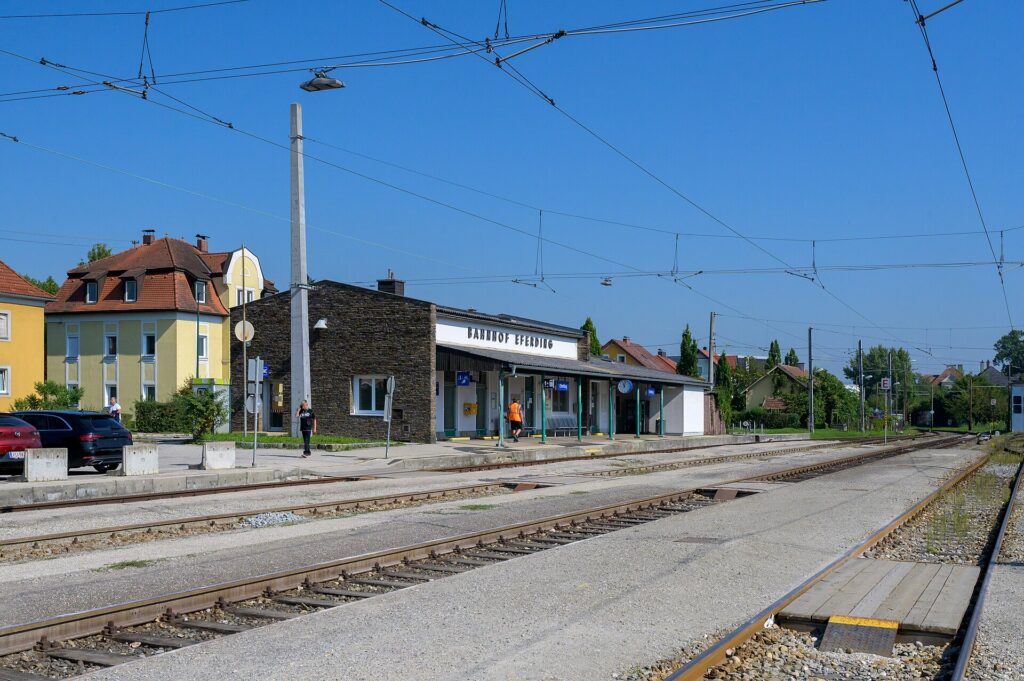 Gleisseite des flachen Aufnahmsgebäudes von Eferding mit dem Schriftzug Bahnhof Eferding und Bruchsteinfassade, davor der überdachte Bahnsteig und die Oberleitung über mehreren Gleisen unter blauem Himmel