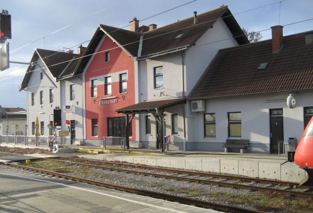 Das Bahnhofsgebäude von Eisenstadt von der Gleisseite: ein weiß-roter Bau mit mehreren Satteldächern und dem Schriftzug EISENSTADT, davor der Bahnsteig mit Bank und Vordach, im Vordergrund Gleise und rechts die Spitze eines roten Triebwagens
