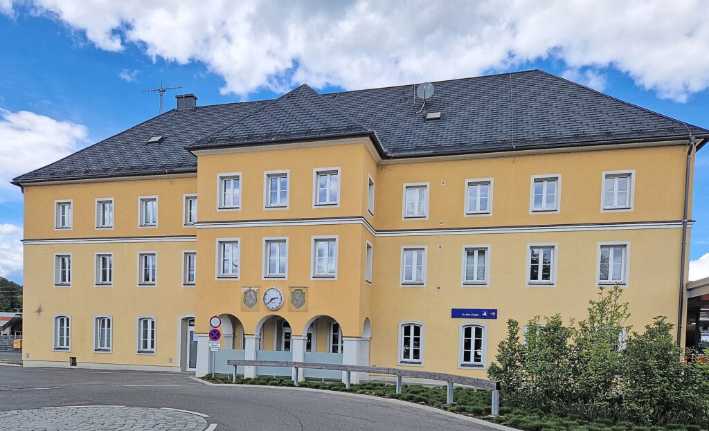Das gelbe, dreigeschoßige Bahnhofsgebäude von Feldbach mit dunklem Walmdach von der Straßenseite, in der Mitte ein Vorbau mit Bahnhofsuhr und drei Rundbögen am Eingang, davor Bäume und Sträucher