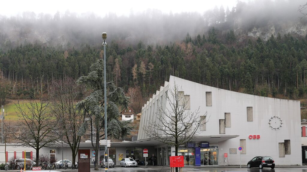 Das helle Bahnhofsgebäude von Feldkirch mit schräg gefalteter Fassade, runder Uhr und ÖBB-Schriftzug, davor der Vorplatz mit parkenden Autos, dahinter ein bewaldeter Hang im Nebel an einem trüben Wintertag