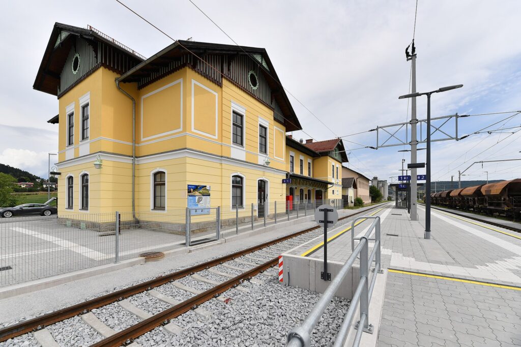 Gelbes, zweigeschoßiges Aufnahmsgebäude des Bahnhofs Freistadt mit holzverzierten Giebeln hinter dem Gleis, rechts der neue Mittelbahnsteig mit Bahnsteigschildern und abgestellten Güterwagen unter bewölktem Himmel