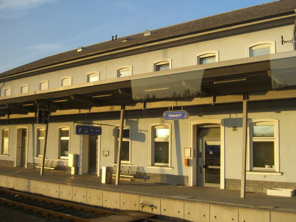 Das helle zweigeschoßige Bahnhofsgebäude von Gleisdorf von der Gleisseite, mit Rundbogenfenstern, einem langen Bahnsteigdach, blauem Stationsschild „Gleisdorf“ und Bänken am Hausbahnsteig im Abendlicht