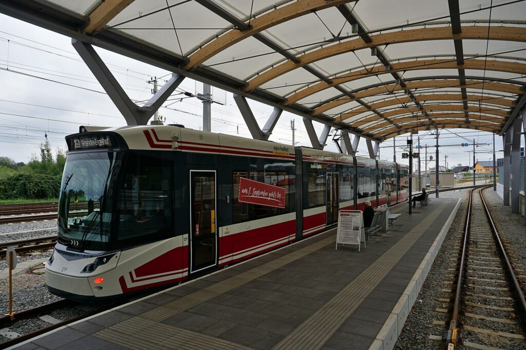 Ein rot-weißer Niederflur-Straßenbahnzug der Traunseetram am Bahnsteig im Bahnhof Gmunden unter einem gewölbten Dach mit Holzbögen und heller Membran, rechts ein leeres Gleis, darüber bewölkter Himmel
