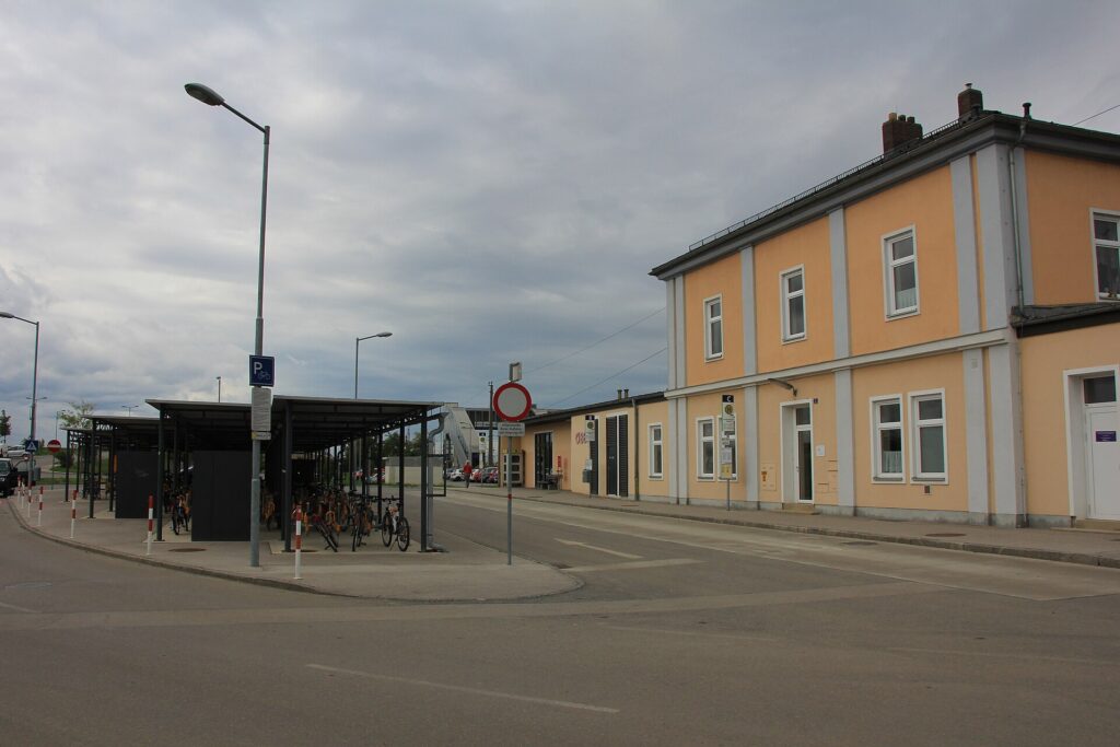 Das zweigeschoßige, apricotfarbene Bahnhofsgebäude von Gramatneusiedl mit weißen Lisenen, links eine überdachte Fahrradabstellanlage, davor eine Straße mit Halteverbotsschild, bewölkter Himmel