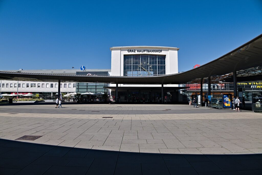 Der Grazer Hauptbahnhof vom Europaplatz aus: ein heller Baukörper mit großer Glasfront, Uhr und dem Schriftzug Graz Hauptbahnhof, davor ein geschwungenes Dach über dem Vorplatz