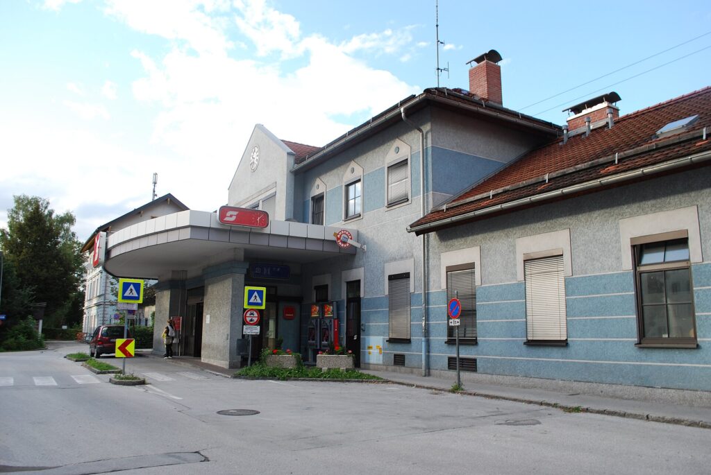 Das Bahnhofsgebäude von Hallein von der Straßenseite: ein zweistöckiger Bau mit hellblau gestreiftem Sockel und Satteldach, davor ein flaches Vordach über dem Eingang mit ÖBB-Logo, links Bäume, vorne die Straße mit Verkehrszeichen
