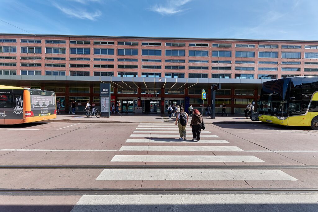 Die lange, rötliche Fassade des Innsbrucker Hauptbahnhofs mit Fensterbändern und einem flachen Vordach über dem Eingang, davor ein Zebrastreifen über den Südtiroler Platz und zwei Busse