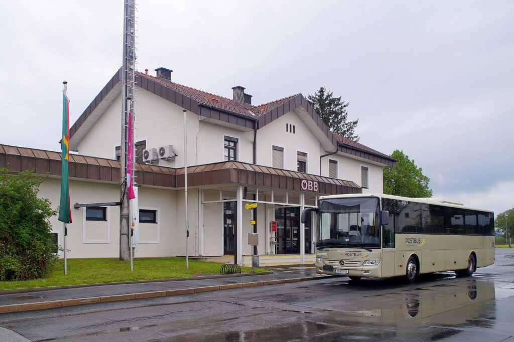 Das zweigeschoßige helle Empfangsgebäude von Jennersdorf von der Straßenseite mit ÖBB-Schriftzug über dem Vordach, davor ein beiger Postbus auf nasser Fahrbahn