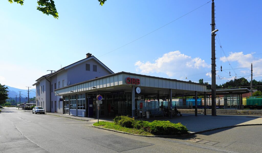 Das zweigeschoßige helle Bahnhofsgebäude von Judenburg mit Satteldach, davor ein flacher Vorbau mit langem Vordach, rotem ÖBB-Logo und Uhr; rechts Güterwagen auf den Gleisen, blauer Himmel