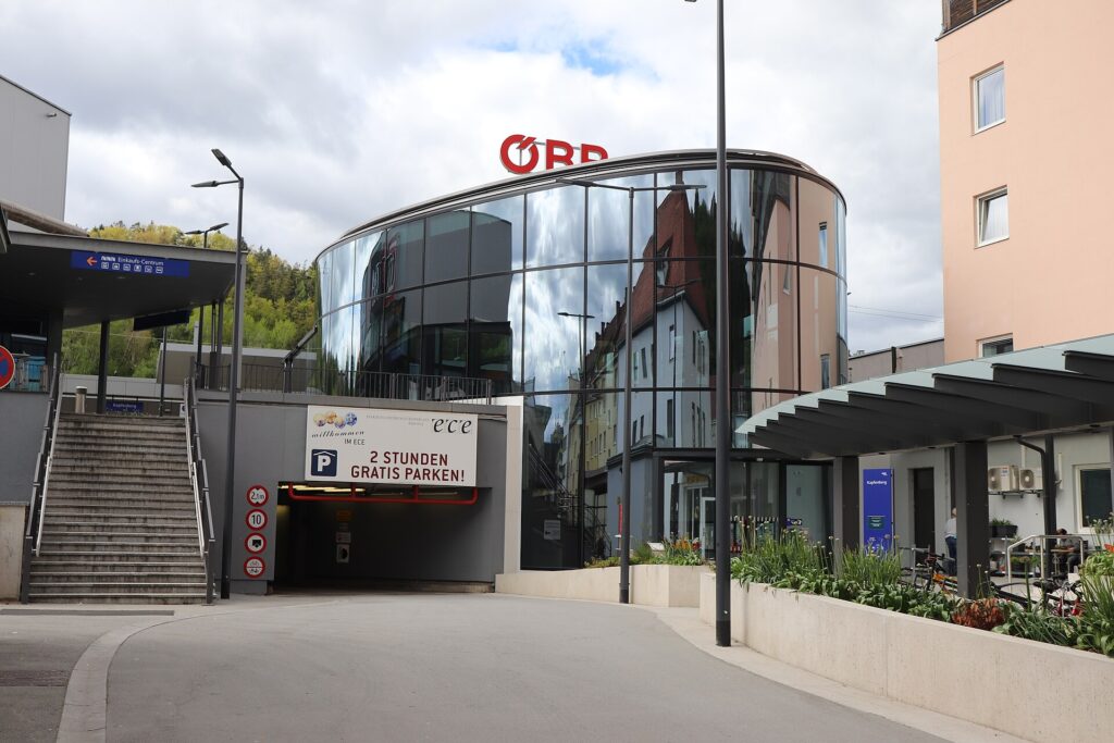Das runde, verglaste Bahnhofsgebäude von Kapfenberg mit ÖBB-Schriftzug auf dem Dach, links eine Treppe und eine Garageneinfahrt, rechts ein überdachter Vorplatz mit Pflanzbeeten