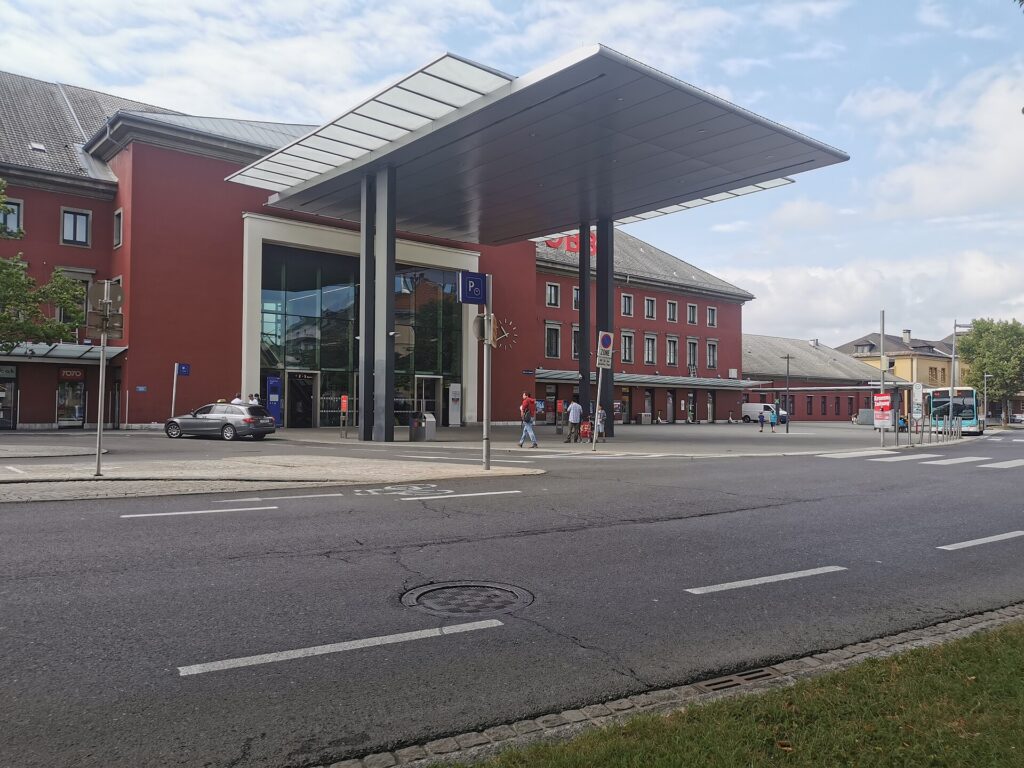 Das rote Bahnhofsgebäude des Klagenfurter Hauptbahnhofs mit verglastem Haupteingang und weit auskragendem Vordach, davor der Vorplatz und Stadtbusse