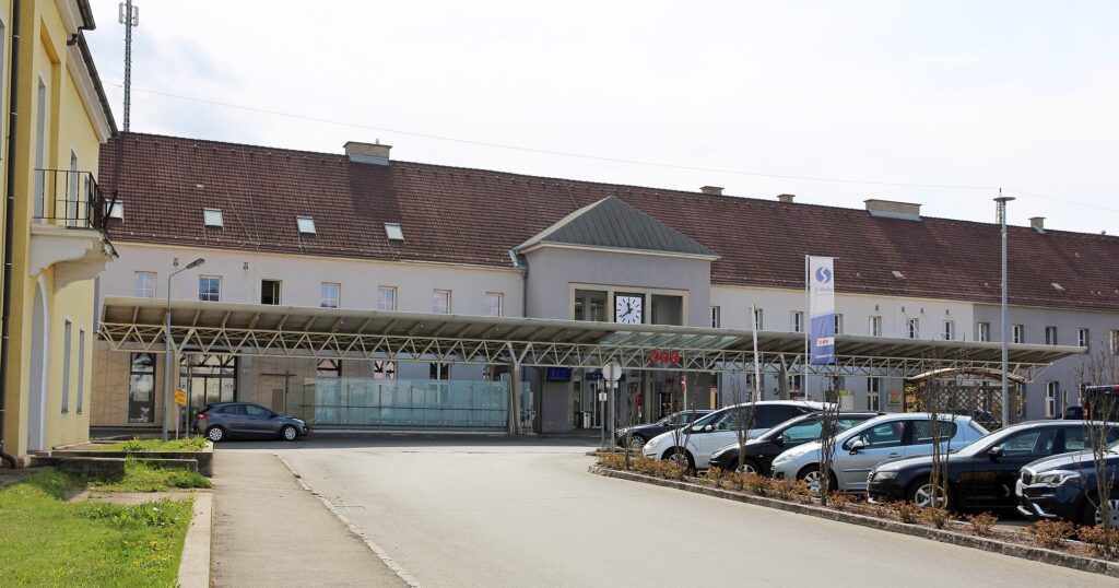 Das lang gestreckte zweigeschoßige Bahnhofsgebäude von Knittelfeld mit rotbraunem Ziegeldach und Uhr im Mittelteil, davor ein durchgehendes Vordach aus Stahl und Glas und parkende Autos am Vorplatz
