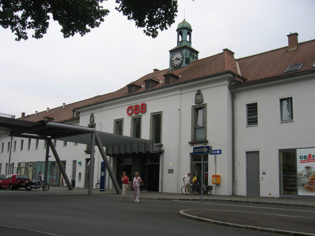 Das hell verputzte Bahnhofsgebäude von Krems mit rotem Ziegeldach, Gauben und grünem Uhrtürmchen, davor ein geschwungenes Stahlvordach und der Vorplatz mit zwei Fußgängerinnen, links parkende Autos
