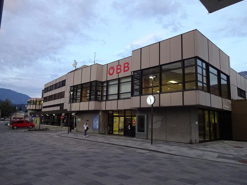 Das zweigeschoßige Bahnhofsgebäude von Kufstein mit heller Plattenfassade, Fensterbändern und rotem ÖBB-Schriftzug in der Abenddämmerung, davor der gepflasterte Vorplatz mit Bahnhofsuhr und einigen Passanten