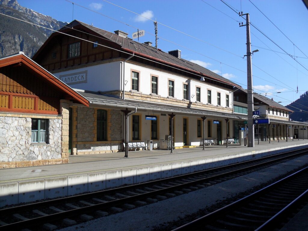 Gleisseitige Ansicht des hellen, zweigeschoßigen Bahnhofsgebäudes mit Steinsockel, Holzgiebel und Schriftzug „LANDECK“, davor der leere Hausbahnsteig mit Vordach, im Hintergrund ein Berghang unter blauem Himmel