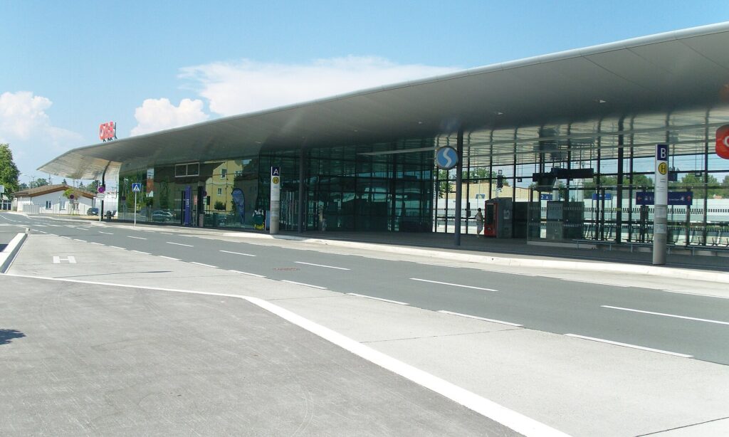 Die verglaste Bahnhofshalle von Leibnitz unter einem lang gestreckten, geschwungenen Flugdach, davor das leere Busterminal mit den Steigmasten A und B unter blauem Himmel