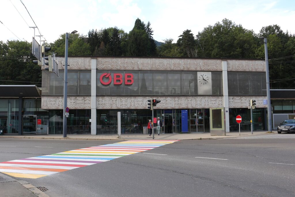 Die flache Front des Leobener Hauptbahnhofs mit Glasfassade, hellen ornamentierten Fassadenbändern, rotem ÖBB-Logo und Uhr, davor eine Straße mit regenbogenfarbenem Zebrastreifen und Ampeln