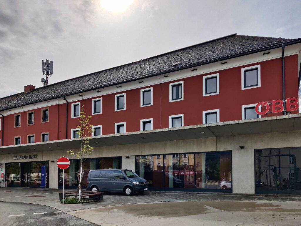 Schrägansicht des dreigeschoßigen, rot verputzten Aufnahmsgebäudes mit weiß gerahmten Fenstern, Walmdach und ÖBB-Logo, darunter das lange Betonvordach mit Glasfront und Schriftzug „Bahnhof Lienz“, davor ein Kleinbus auf dem Vorplatz