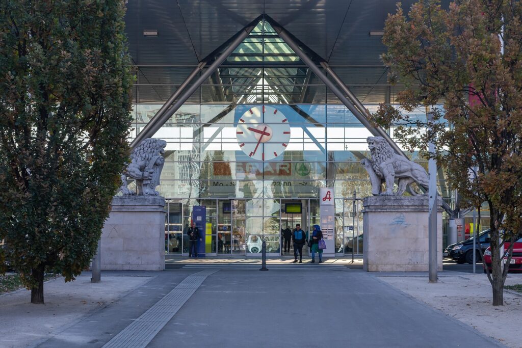 Der Haupteingang des Linzer Hauptbahnhofs: eine hohe Glasfassade mit großer Bahnhofsuhr unter einem Dach mit schrägen Stahlstreben, links und rechts des Eingangs je ein Steinlöwe auf einem hohen Sockel, davor ein Gehweg zwischen zwei Bäumen