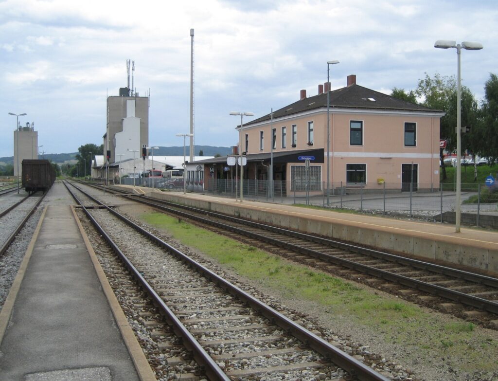 Blick entlang der Gleise auf das zweistöckige, hellrosa Empfangsgebäude mit Walmdach und eingeschoßigem Vorbau am Bahnsteig, links ein abgestellter Güterwagen und Silotürme