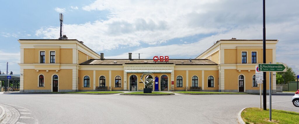 Frontansicht des gelben, symmetrischen Aufnahmsgebäudes von Melk mit zwei zweigeschoßigen Eckbauten und niedrigem Mitteltrakt mit Vordach und ÖBB-Schriftzug, davor ein leerer Vorplatz und eine Skulptur, darüber blauer Himmel mit Wolken