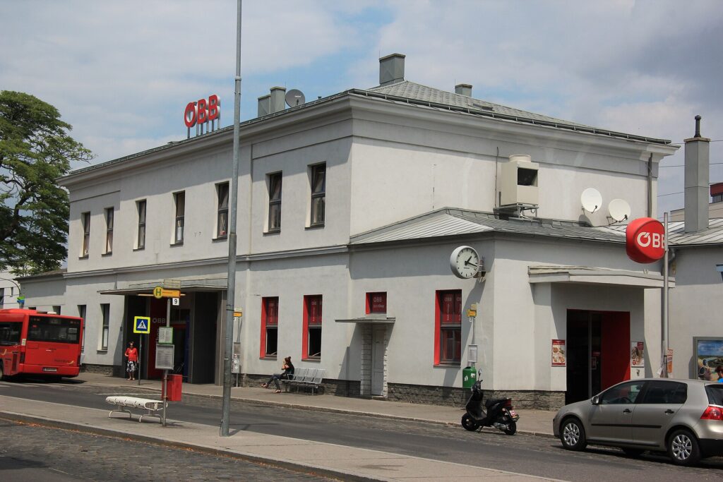 Das hellgraue zweigeschoßige Bahnhofsgebäude von Mödling mit ÖBB-Logos, Bahnhofsuhr und rot gerahmten Fenstern, davor ein Gehsteig mit Bank, links ein roter Bus, rechts ein geparktes Auto