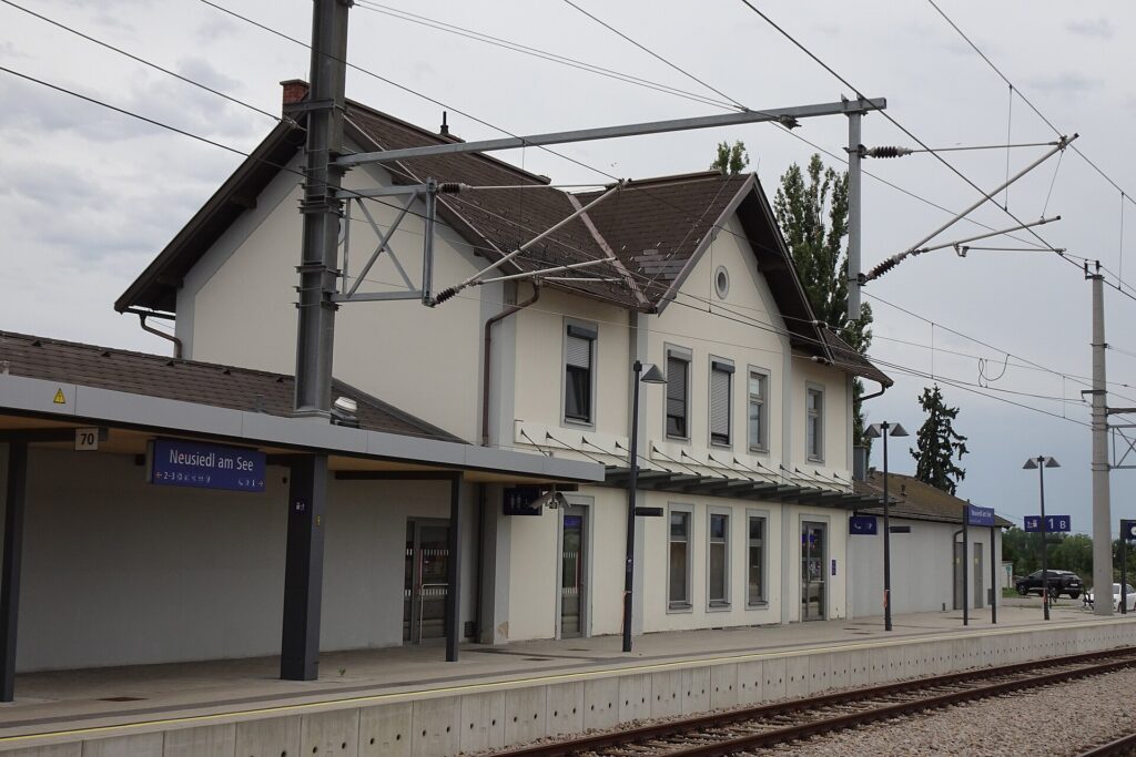 Das zweigeschoßige, hell verputzte Aufnahmsgebäude mit Satteldächern von der Gleisseite aus gesehen, davor das überdachte Bahnsteigende mit dem Stationsschild Neusiedl am See und die Oberleitung
