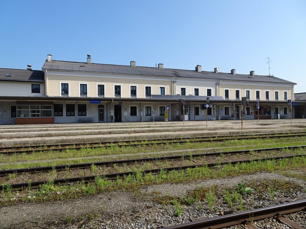 Blick über mehrere Gleise auf das lange, zweigeschoßige hellgelbe Aufnahmsgebäude von Ried im Innkreis mit Bahnsteigdach unter blauem Himmel