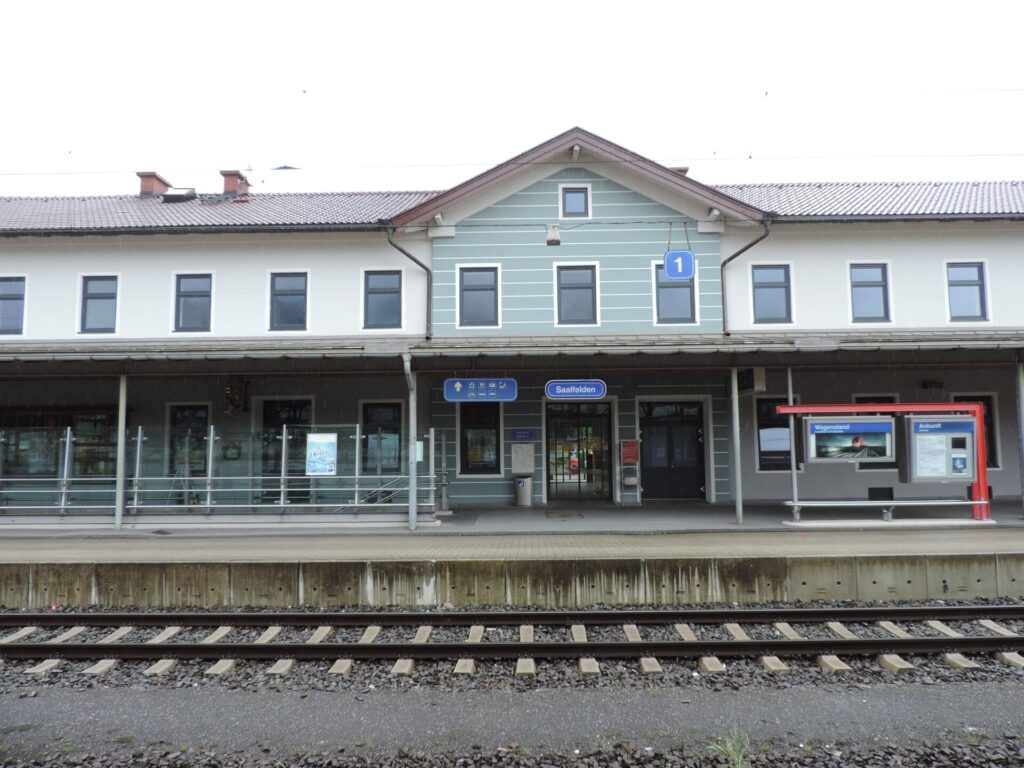 Das zweistöckige Bahnhofsgebäude von Saalfelden von der Gleisseite: ein heller Bau mit grau-grün gestreiftem Mittelteil unter einem Giebel, davor das Bahnsteigdach mit dem Schild Saalfelden und im Vordergrund das Gleis 1