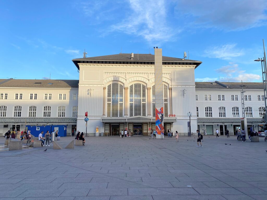 Das helle, denkmalgeschützte Empfangsgebäude des Salzburger Hauptbahnhofs mit großen Bogenfenstern und ÖBB-Logo, davor der weite, gepflasterte Südtiroler Platz