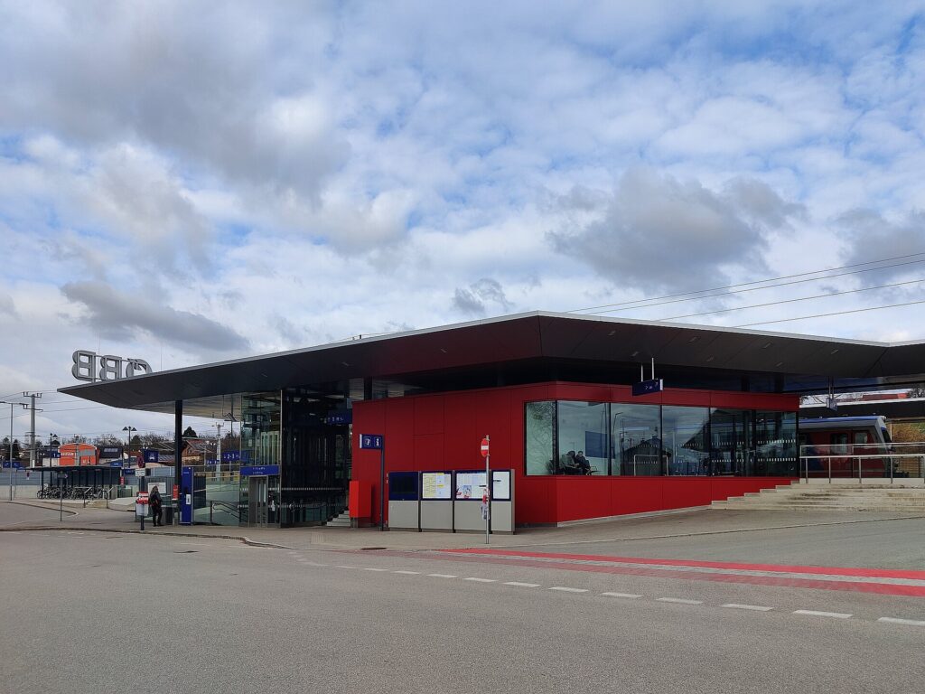 Rot verkleideter, verglaster Warteraum des Bahnhofs Schärding unter einem weit auskragenden dunklen Flachdach mit ÖBB-Schriftzug, links der Abgang zur Unterführung mit Aufzug und abgestellte Fahrräder, davor der Bus-Vorplatz unter bewölktem Himmel