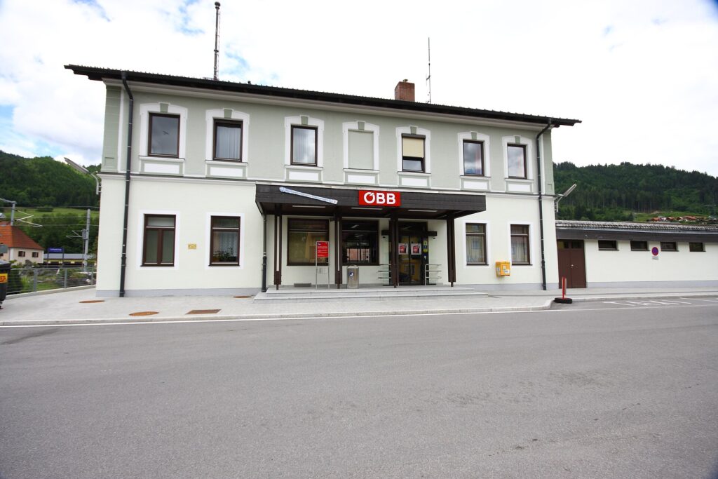 Das zweigeschoßige hellgrüne Bahnhofsgebäude von Schladming mit weißen Fensterumrahmungen und ÖBB-Schriftzug über dem dunklen Vordach, von der Straße aus gesehen, dahinter bewaldete Hänge