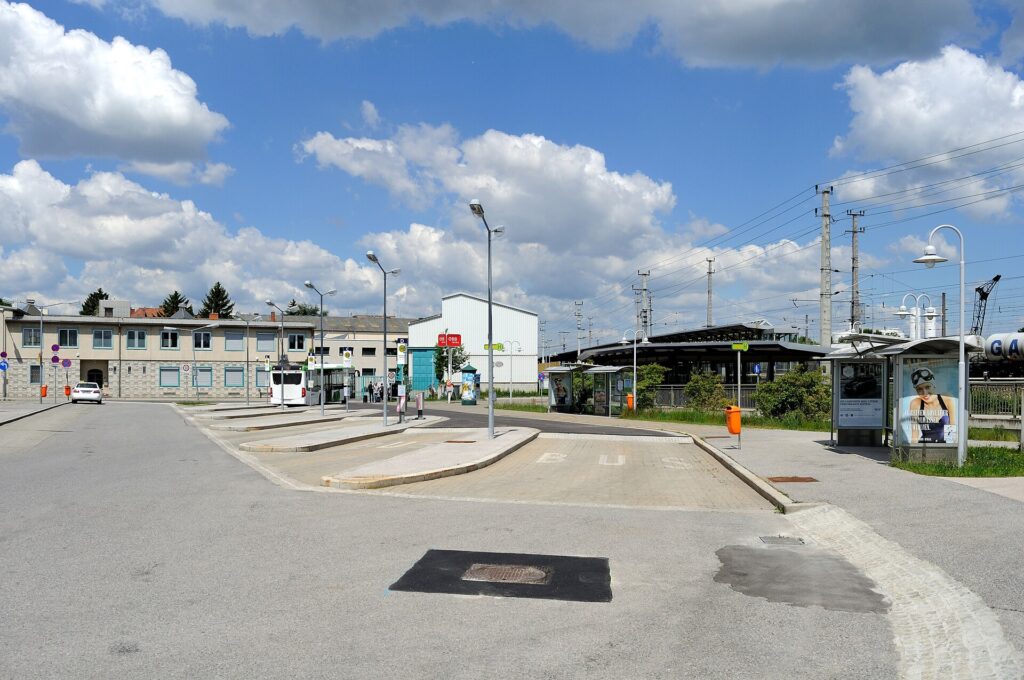 Der leere Busbahnhof Schwechat mit Bussteiginseln und einem Wartehäuschen, im Hintergrund ein zweigeschoßiges Gebäude, eine weiße Halle und das Bahnsteigdach mit Oberleitungsmasten unter blauem Himmel mit Wolken