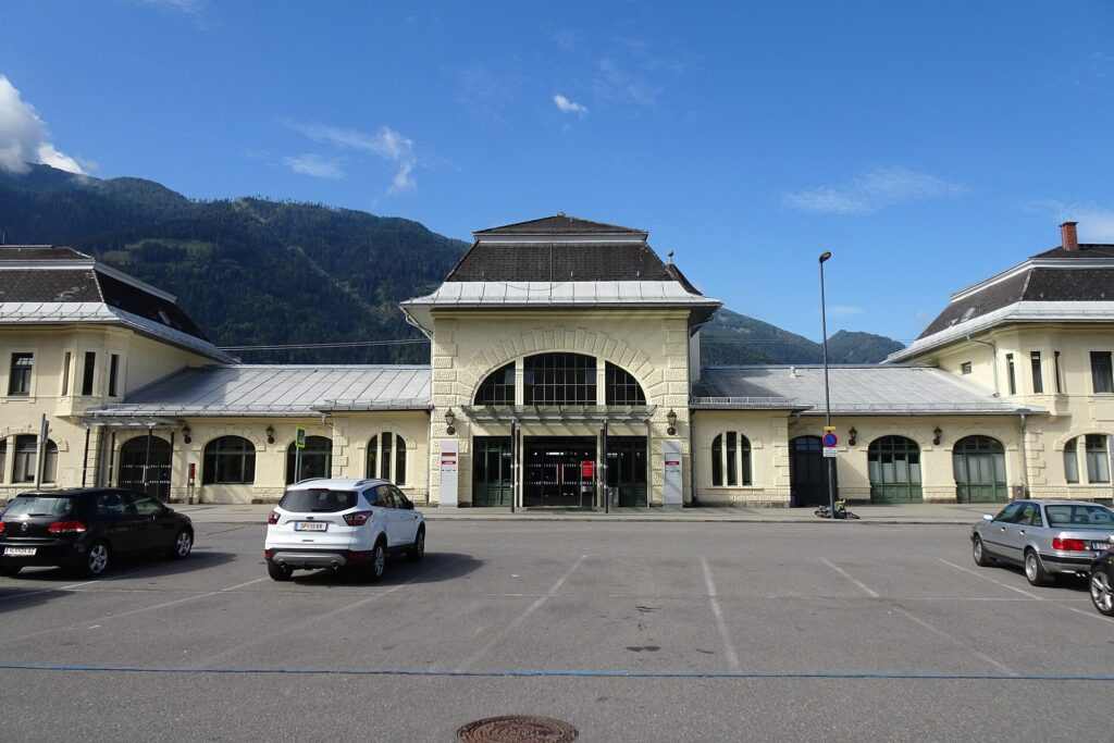 Frontansicht des hellgelben Aufnahmsgebäudes von Spittal-Millstättersee mit hohem Mittelbau, großem Rundbogenfenster und Mansarddach, eingeschoßigen Verbindungstrakten und zwei Seitenflügeln, davor der Parkplatz mit einigen Autos und bewaldete Berge