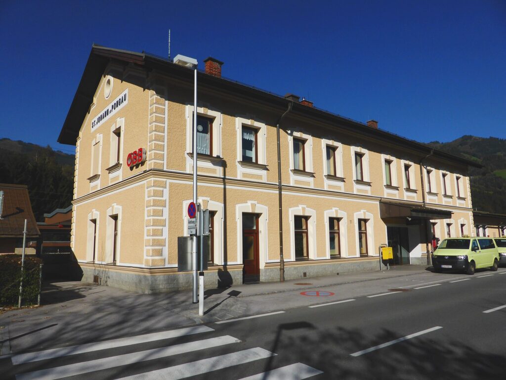 Schrägansicht des zweigeschoßigen, ockerfarbenen Aufnahmsgebäudes von St. Johann im Pongau mit weißen Fensterumrahmungen, dem Schriftzug St. Johann i. Pongau am Giebel und ÖBB-Logo, davor die Straße mit Zebrastreifen und zwei Taxis unter blauem Himmel