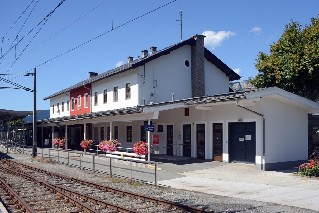 Gleisseitige Schrägansicht des zweigeschoßigen weißen Aufnahmsgebäudes mit rot abgesetztem Gebäudeteil und eingeschoßigem Vorbau mit Vordach, davor Blumentröge am Bahnsteig und Gleise im Vordergrund