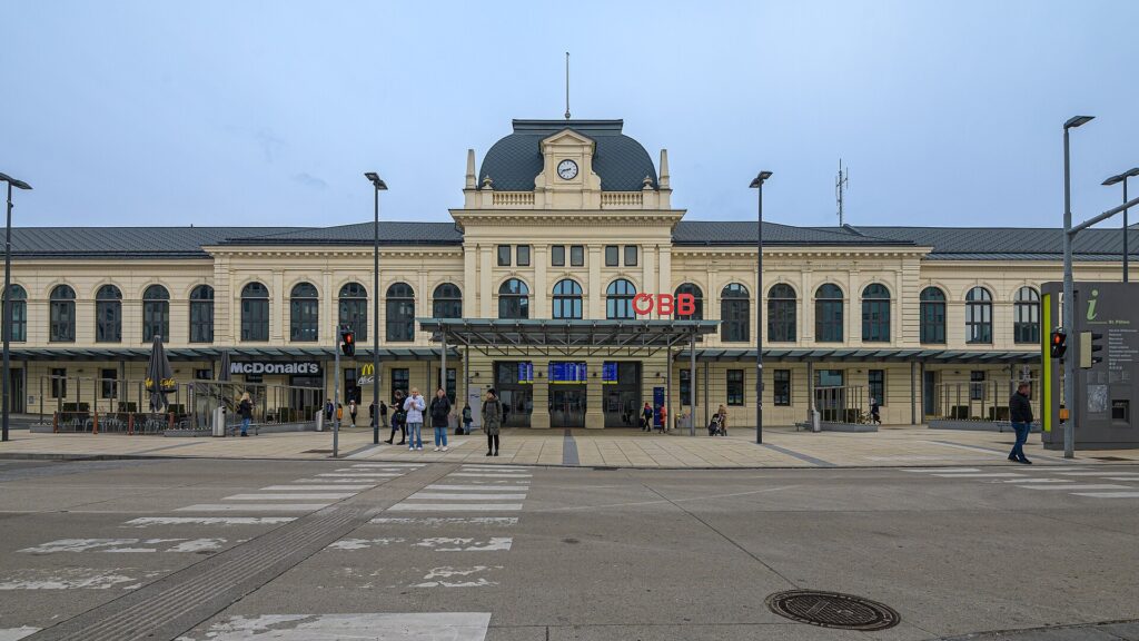 Frontalansicht des cremefarbenen historischen Bahnhofsgebäudes von St. Pölten mit Uhrturm und Kuppeldach, Rundbogenfenstern, ÖBB-Logo und gläsernem Vordach über dem Eingang, davor der Bahnhofplatz mit Zebrastreifen